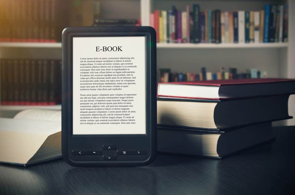 KDP book listing optimization: A Kindle eBook reader stands on a desk with textbooks and a bookshelf in the background.